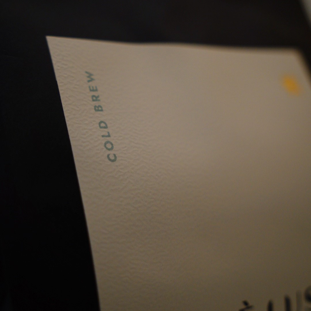 A close-up of a textured off-white paper label with the words 'COLD BREW' printed vertically in dark green, set against a dark background.