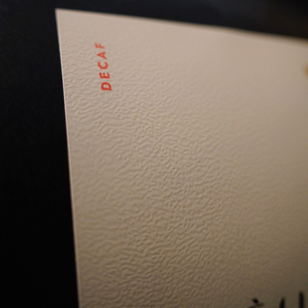 Close-up of a textured cream-colored label with the word 'DECAF' printed vertically in red near the top left, set against a dark background.