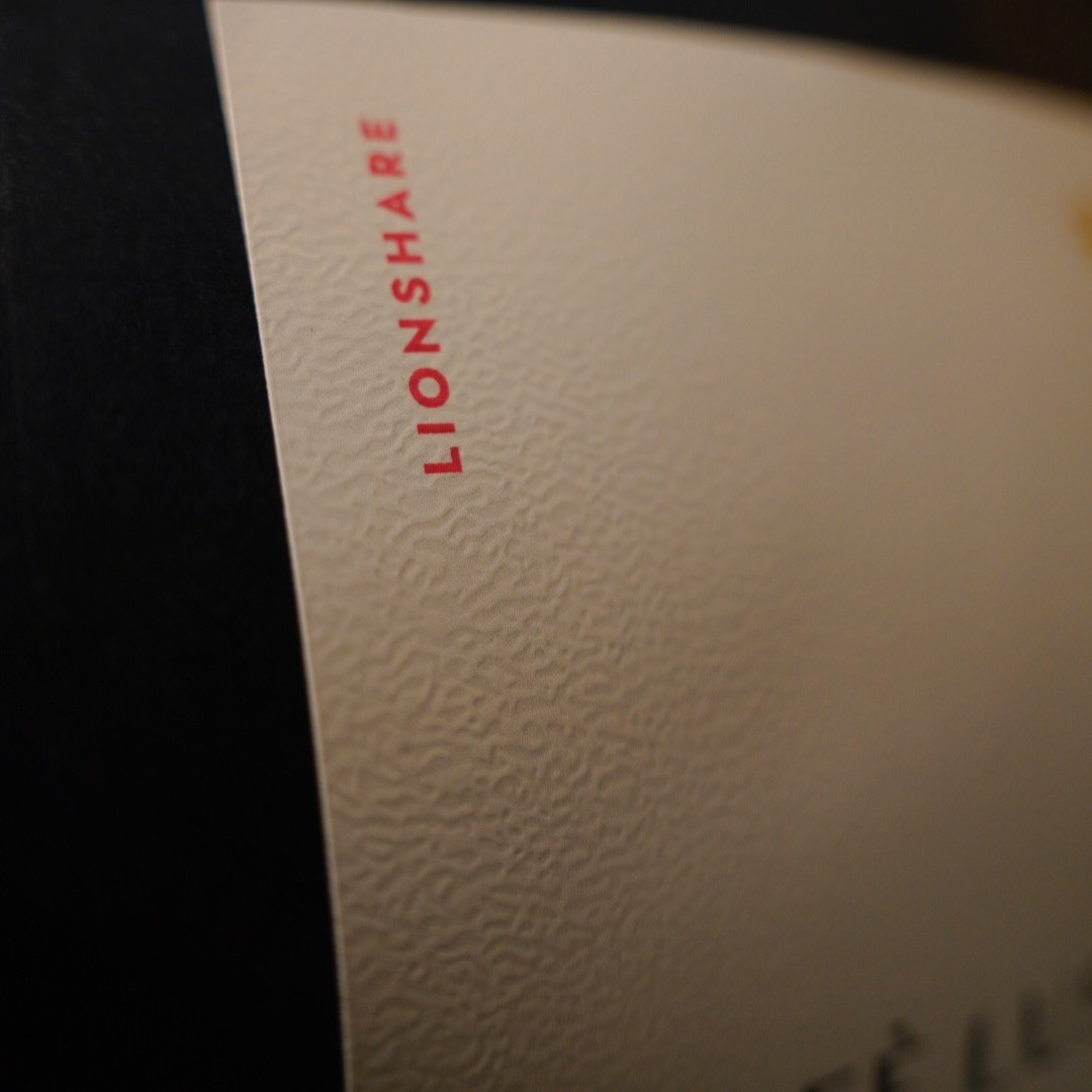 A close-up of a textured off-white label with the word 'LIONSHARE' printed vertically in red, next to a dark black background.