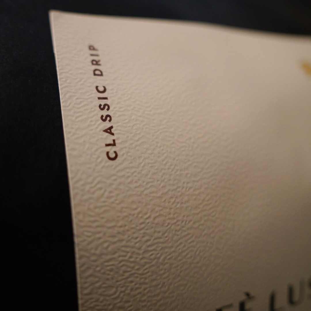 Close-up of a cream-colored textured label corner with the words 'CLASSIC DRIP' printed vertically in dark brown, set against a dark background.