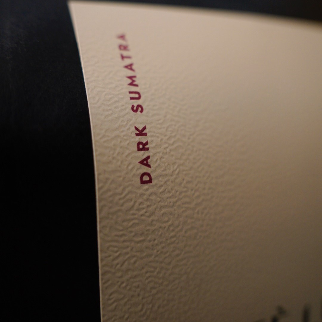 Close-up of a textured cream-colored  label with the words 'DARK SUMATRA' printed vertically in dark red, next to a black background.