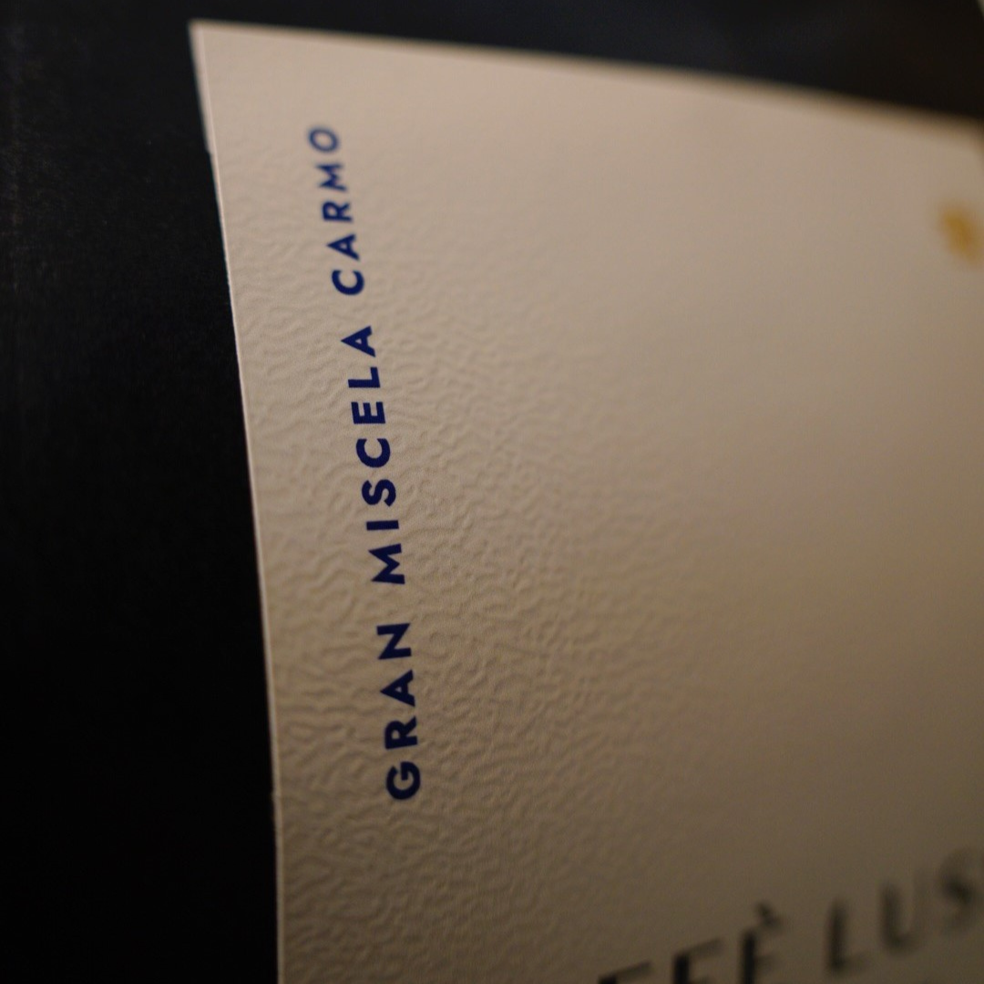 Close-up of a cream-colored textured label with dark navy blue text reading 'GRAN MISCELA CARMO' on a dark background.