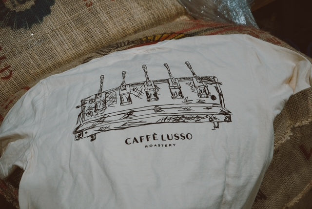 A person wearing a white crew neck t-shirt with a printed design of espresso levers and the text 'CAFFÈ LUSSO' on the front, sitting in a setting with coffee storage in the background.