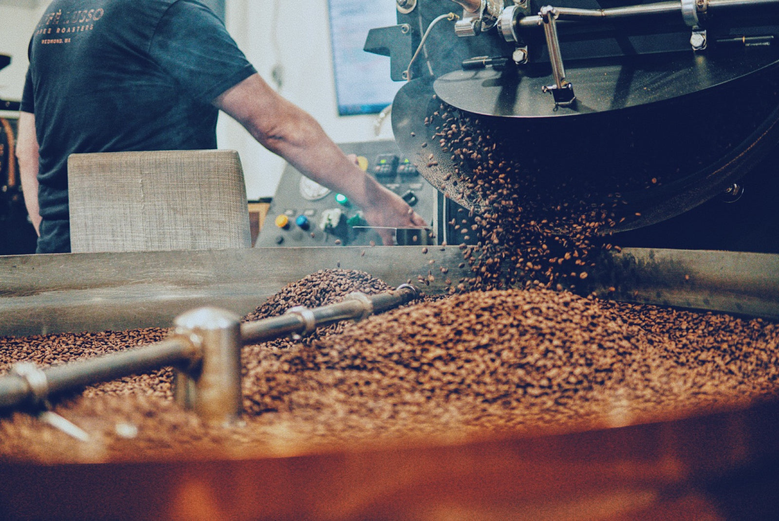 COffee dropping out of a large coffee roasting machine while the roaster controls the drop inthe background