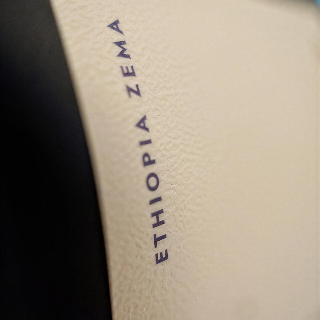 Close-up of a textured cream-colored leather or faux leather surface with the brand name 'ETHIOPIA ZEMA' printed vertically in dark navy blue. A black curved shape is visible on the left side of the frame.