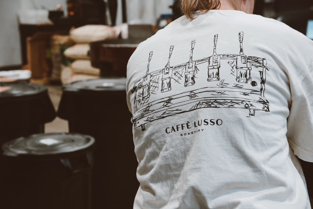 A person wearing a white crew neck t-shirt with a printed design of espresso levers and the text 'CAFFÈ LUSSO' on the front, sitting in a setting with coffee storage in the background.