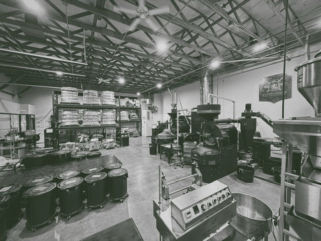 a wide anlge view of the roastery in redmond, WA. Stacks of coffee sacks, barrels of stored beans and two coffee roasting machines.